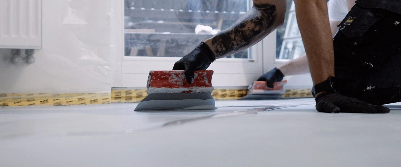 Ręczna aplikacja mikrocementu: dwie osoby prowadzą metalowe pace po podłodze.
